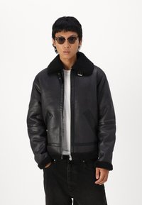 Schwarze Lederbomberjacke mit einem Schaffellkragen und -manschetten, ausgestattet mit einem Reißverschluss vorne und Seitentaschen. Kombiniert mit einem weißen T-Shirt und dunklen Jeans.
