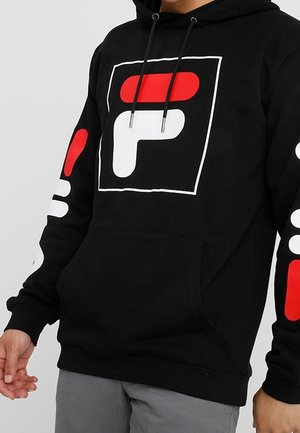 Schwarzer Hoodie mit großem weißem und rotem geometrischem Logo auf Brust und Armen, getragen mit grauer Hose, Hände und Mittelteil sichtbar.