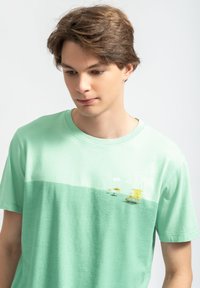 Mintgroene t-shirt met korte mouwen, featuring een gradient ontwerp, met gele lifeguard hutten en parasols gedrukt op de voorkant. Zachte stoftextuur.