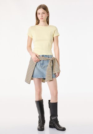 Młoda kobieta w żółtym topie z krótkim rękawem, jeansowej spódnicy, czarnych kozakach za kolano i w paskowanym swetrze przewiązanym w pasie, stojąca na białym tle.