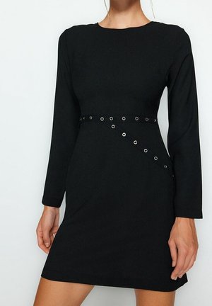 Robe noire à manches longues avec un col rond et une ceinture diagonale à œillets à la taille, portée par un mannequin visible du cou jusqu'à la mi-cuisse.