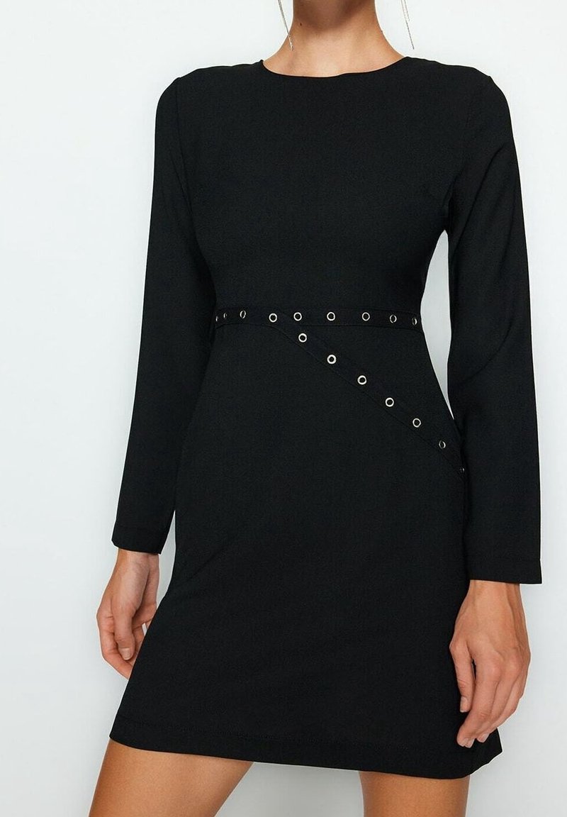 Robe noire à manches longues avec un col rond et une ceinture diagonale à œillets à la taille, portée par un mannequin visible du cou jusqu'à la mi-cuisse.