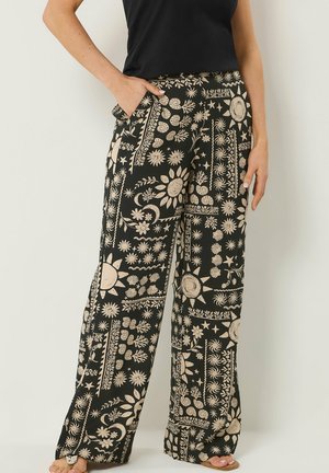 Frau trägt schwarze Hosen mit weitem Bein, verziert mit beigen Mustern von Sonne, Sternen, Mond und Blumen, kombiniert mit einem schwarzen ärmellosen Oberteil.