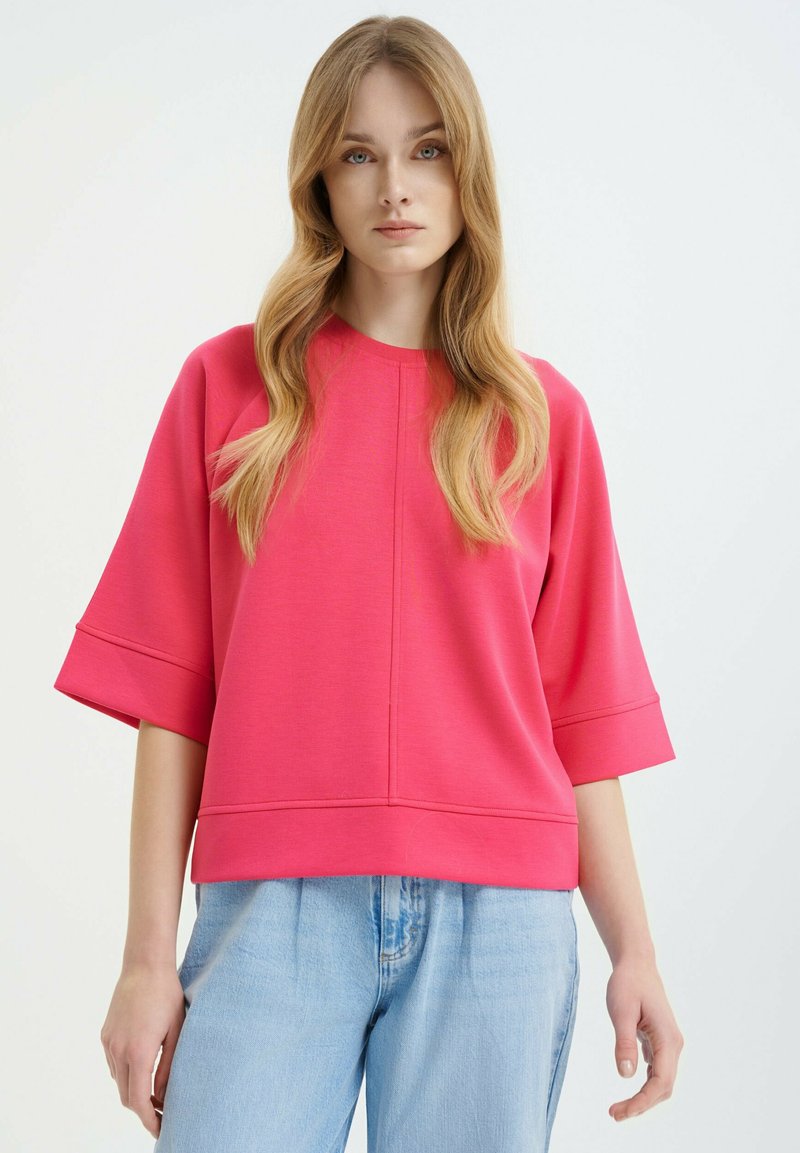Pink sweatshirt with a round neckline, three-quarter sleeves, and a relaxed fit. Made from a soft, textured fabric. Pairing with light blue jeans.