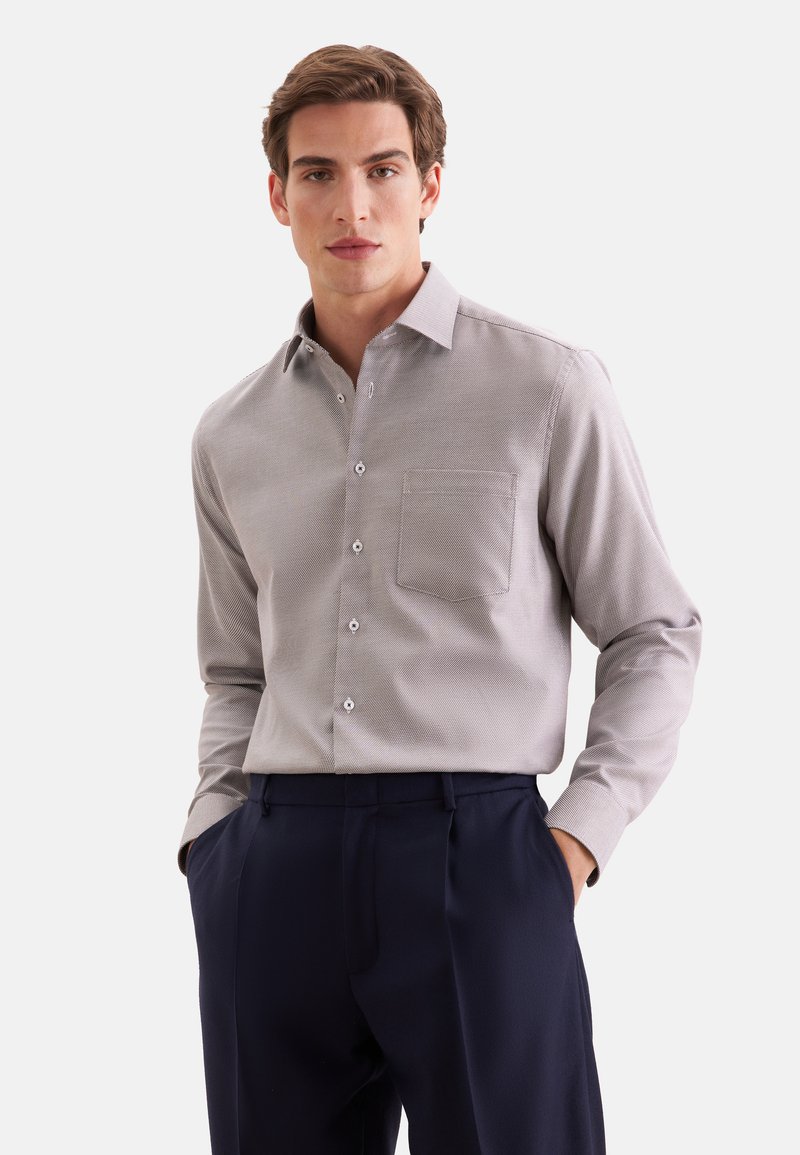 Jeune homme aux cheveux bruns porte une chemise gris clair boutonnée et un pantalon bleu foncé, debout les mains dans les poches devant un fond blanc.