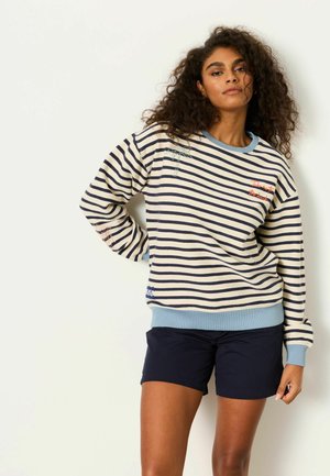 Frau mit lockigem Haar, die einen blau-weiß gestreiften Pullover mit hellblauen Bündchen und marineblaue Shorts trägt, stehend vor einem weißen Hintergrund.