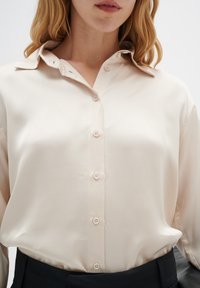 Helle beige Bluse mit spitzem Kragen, ausgestattet mit einer knöpfbaren Vorderseite und glatter Satinstoffstruktur. Lange Ärmel und lockere Passform.