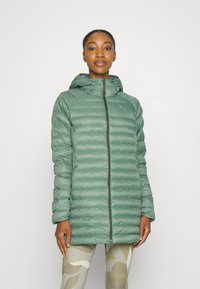 Hellgrüne Pufferjacke mit Kapuze, mit gestepptem Design, durchgehendem Reißverschluss und Logo-Detail. Getragen über gemusterten Leggings.