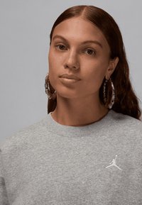 Grå genser med rund hals og ribbet mansjetter, med en hvit Jordan-logo på brystet. Modellen har store hoop-øreringer og bølgete hår.