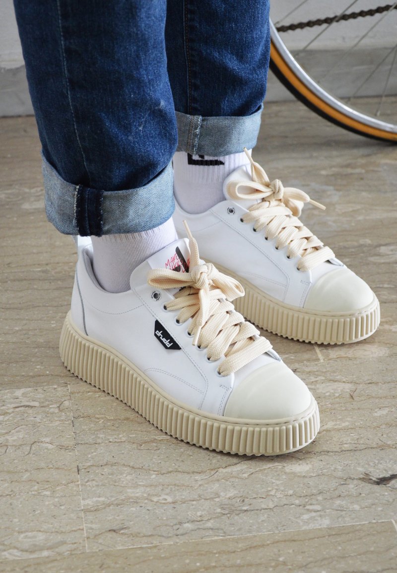 White leather sneakers with a ribbed cream sole, beige laces, and logo tag. Worn with rolled blue denim, cream socks. Smooth texture.