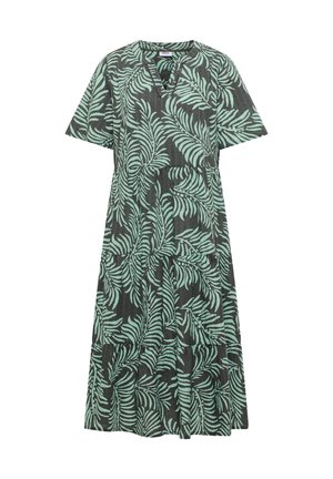 Femme aux cheveux courts et bruns portant une robe verte avec des motifs de feuilles bleu clair, debout, de dos sur un fond beige.
