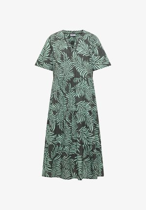 Femme aux cheveux courts et bruns portant une robe verte avec des motifs de feuilles bleu clair, debout, de dos sur un fond beige.