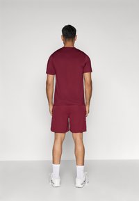 Bordový športový krátky rukáv, ktorý je skombinovaný s ladnými šortkami. Oblečenie má hladkú, ľahkú textúru a nenápadné akcenty loga.