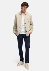 Beige knappestoffjakke med brystlomme, båret over en hvit t-skjorte, kombinert med marineblå bukser og beige joggesko. Glatt materiale, uformell design.