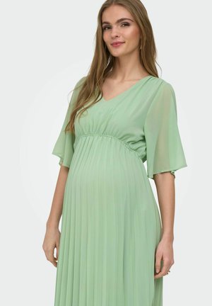 Tehotná žena sa usmieva, nosiaca svetlozelené plisované tehotenské šaty s rozstrapatenými rukávmi, stojaca pred jednoduchým pozadím.