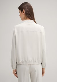 Lichtgrijze jas met een ontspannen pasvorm, elastische cuffs en een knoopdetail op de achterkant. Gemaakt van gladde stof met een eenvoudig ontwerp.