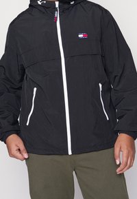 Czarna lekka kurtka z kapturem, białymi detalami na zamku, elastycznymi mankietami oraz naszywką z logo z przodu. Posiada dwie boczne kieszenie.