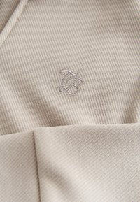 Beiges, geripptes Textil mit subtiler Textur, mit einem kleinen, hellvioletten gestickten Logo. Glatter Stoff mit einem aufgefalteten Kanten-Detail.