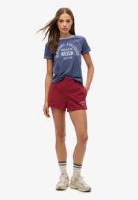 Conjunto informal que incluye una camiseta de algodón azul con estampado blanco y pantalones cortos color burdeos. Los accesorios incluyen calcetines a rayas blancas y azules y zapatillas ligeras.
