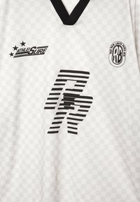 Hvid sports trøje med subtilt ternet mønster, sort V-hals og sorte logos, herunder "AthleiSure" og "RB Test New Limits Paris 1978".