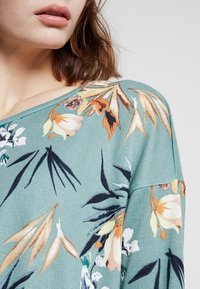 Helles türkisfarbenes Sweater mit floralem Muster in sanften Farben. Er hat einen weiten Halsausschnitt und ein texturiertes Material, das die detaillierten Blumendesigns betont.