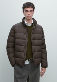 Giacca a vento marrone con colletto alto, design trapuntato, bottoni frontali e texture liscia, indossata sopra un maglione scuro e una camicia bianca.