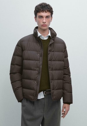 Doudoune marron avec un col haut, un design matelassé, des boutons à l'avant et une texture lisse, portée sur un pull foncé et une chemise blanche.