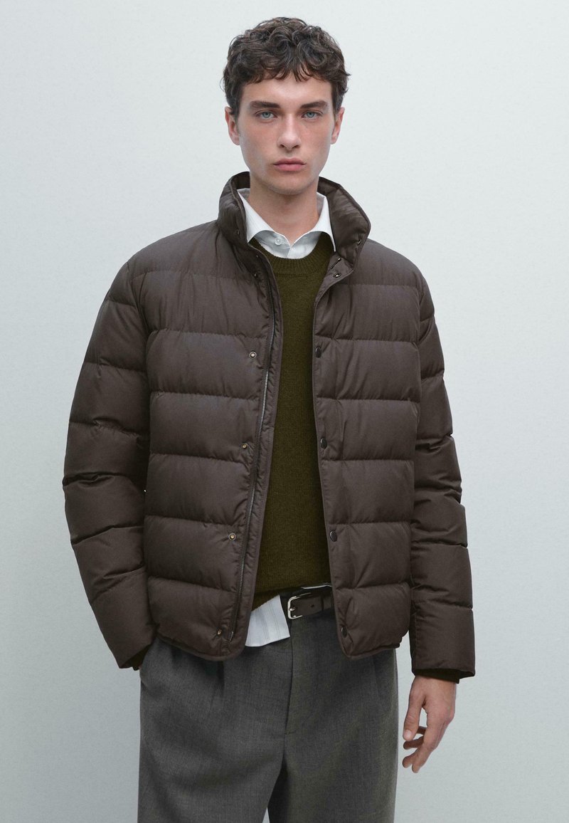 Giacca a vento marrone con colletto alto, design trapuntato, bottoni frontali e texture liscia, indossata sopra un maglione scuro e una camicia bianca.