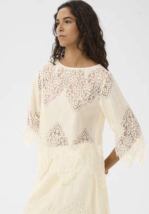 Mujer con un vestido blanco roto con intrincados patrones de encaje floral en el pecho, las mangas y el dobladillo, mirando ligeramente hacia un lado contra un fondo liso.