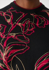 Black knit sweater featuring bold red floral embroidery and gold outlines. Round neckline with a contrasting red trim. Close-up detail.