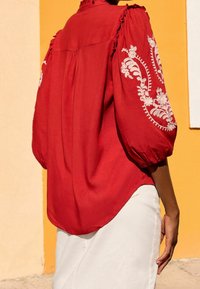 Röd blus med uppblåsta ärmar och vit blommig broderi. Mjukt tyg, rundad nederkant och volangdetaljer vid halsringningen. Bärs med vita byxor.