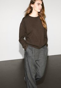 Pull marron à coupe décontractée, col rond et ourlet côtelé, associé à un pantalon en lin léger gris clair avec des plis.