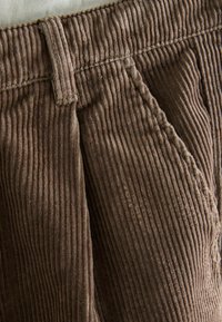 Brown corduroy pants featuring vertical ribbing, a zipper fly, and pleated front. Durable texture with a loose fit.