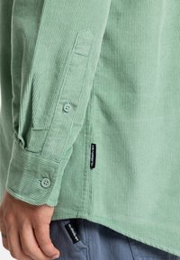 Lichtgroene corduroy shirt met ribbelstructuur, knoopmanchetten en merklabels. Heeft een rechte zoom en een iets langere achterkant.
