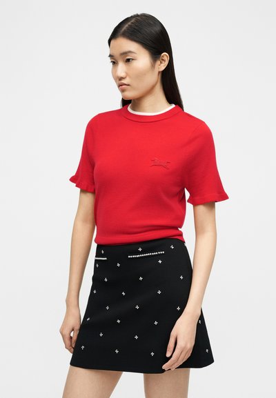 Γυναίκα με ίσια μαύρα μαλλιά φοράει κόκκινη μπλούζα με κοντά μανίκια και κεντημένο λογότυπο, καθώς και μαύρη φούστα με μικρές λευκές διακοσμήσεις.