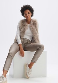 Faux-Fur-Weste in hellbeige, kombiniert mit einem weißen Langarmshirt und beigen, figurbetonten Hosen. Das Model trägt cremefarbene Sneaker mit Akzenten.