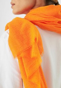 Écharpe légère orange vif drapée sur un haut blanc, avec des bords effilochés et une texture douce, mettant en valeur un design ample et décontracté.