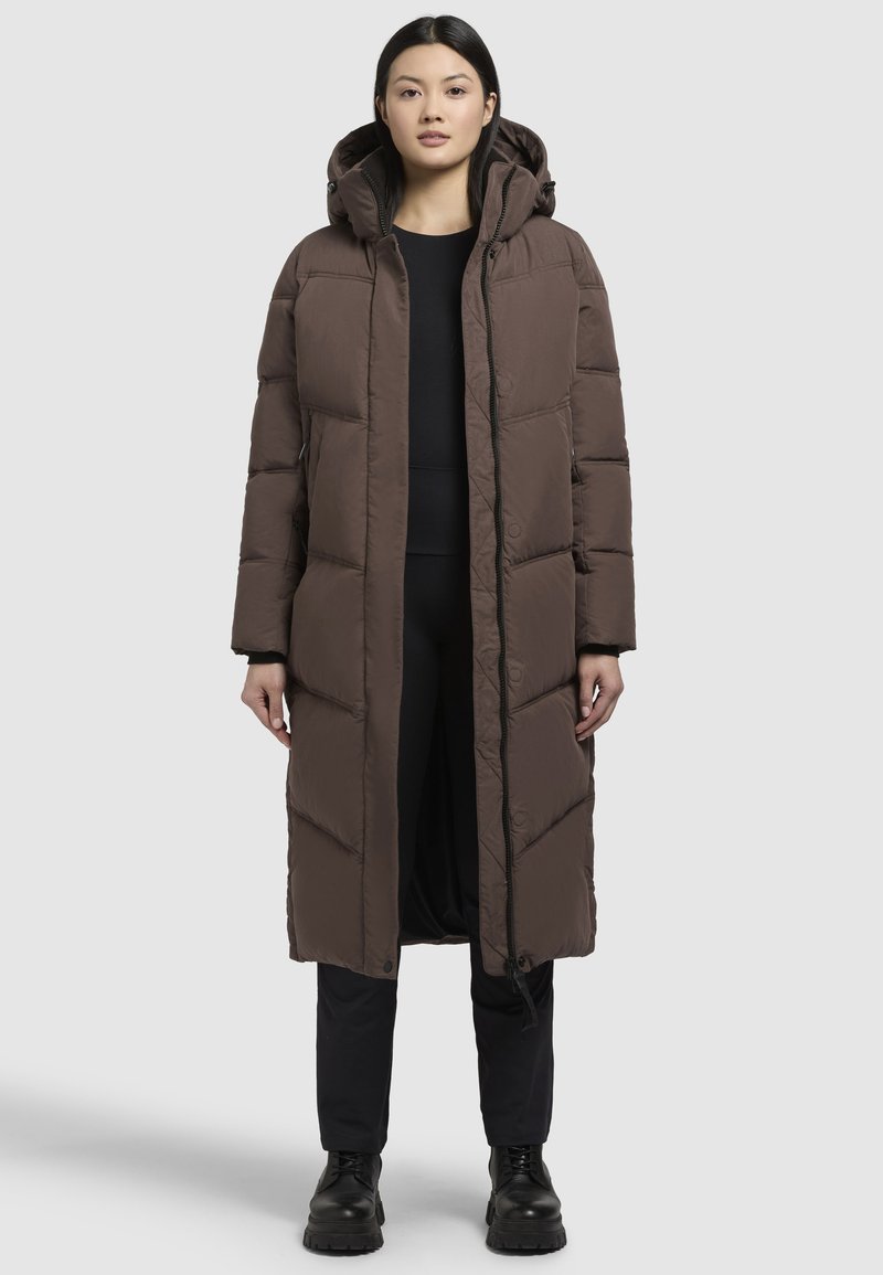Langer brauner Puffer-Mantel mit Kapuze, ausgestattet mit einer gew-quilteten Textur und einem Frontreißverschluss. Trägt einen schwarzen Look und passt zu schwarzen Stiefeln.