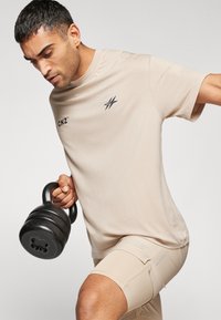 Beež sportlik t-särk lühikeste varrukatega, millel on väike tumesinine logo, ja sobivad beežid lühikesed püksid, hoides musta kettlebelli.