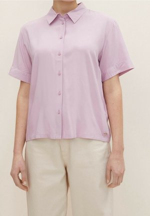 Koszula na guziki o krótkim rękawie w kolorze liliowym, noszona z kremowymi spodniami z wysokim stanem na stojącej osobie, neutralne tło.