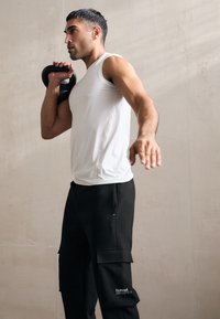 Balta be rankovių sportinė marškinėliai, derinami su juodomis krovininėmis kelnėmis, turinčiomis kišenes ir logotipą, laikantis kettlebell, neutralioje fone.