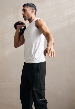 Weißes ärmelloses Sportshirt kombiniert mit schwarzen Cargo-Hosen mit Taschen und Logo, hält eine Kettlebell, vor einem neutralen Hintergrund.