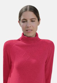 Pull en tricot de couleur rose vif, avec un col montant, un motif en losanges et des poignets côtelés. Tissu doux et texturé avec une silhouette ajustée.