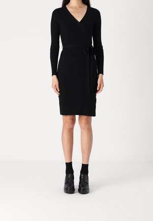 Femme portant une robe portefeuille en maille côtelée noire à manches longues, une ceinture à la taille, des chaussettes noires aux chevilles et des chaussures plateformes noires, debout sur un fond blanc.