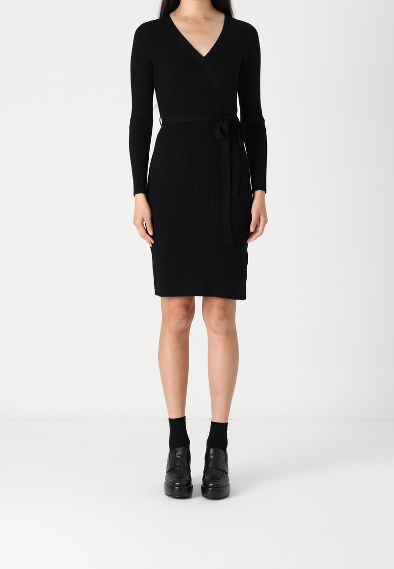 Femme portant une robe portefeuille en maille côtelée noire à manches longues, une ceinture à la taille, des chaussettes noires aux chevilles et des chaussures plateformes noires, debout sur un fond blanc.