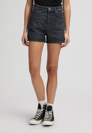 Άτομο που φοράει μαύρα τζιν σορτς, μαύρο τοπ, λευκές κάλτσες και μαύρα ψηλά αθλητικά παπούτσια, στέκεται μπροστά σε απλό φωτεινό φόντο.