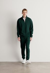 Completo sportivo verde scuro composto da un pullover con zip e colletto, abbinato a pantaloni affusolati. Il materiale sembra morbido, con una consistenza liscia.