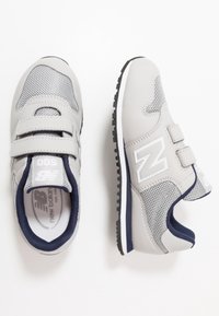 Grå sportskor med meshpaneler, kardborreband och marinblå detaljer. Har en vit logotyp och ett mönstrat gummisula för bra grepp.