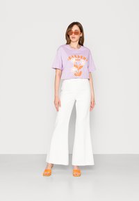T-shirt lavanda com gráfico de flor laranja, emparelhado com calças brancas de boca de sino e sandálias de salto laranja, colocado contra um fundo neutro.