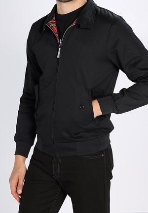 Mann trägt eine schwarze Jacke mit Reißverschluss und rotem Karomuster am inneren Kragen, die Hände in den Taschen, gekleidet in schwarze Hose vor einfachem Hintergrund.